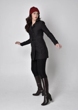 Siyah ceketli, deri çizmeli ve kırmızı bereli güzel esmer bir kızın tam boy portresi. Gri bir stüdyonun arka planında dikiliyor..