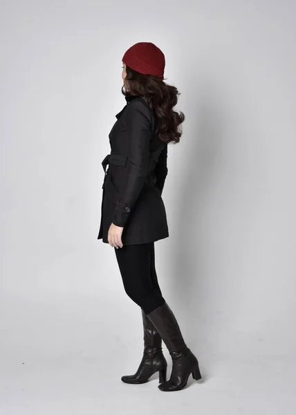 黒のコート 革のブーツと赤いビーニーを身に着けているかなりブルネットの女の子の完全な長さの肖像画 灰色のスタジオの背景に立つポーズ — ストック写真