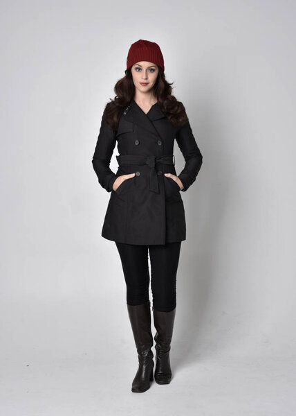 портрет красивой брюнетки в черном пальто, кожаных сапогах и красной шапочке. Стоящая поза на сером студийном фоне
.