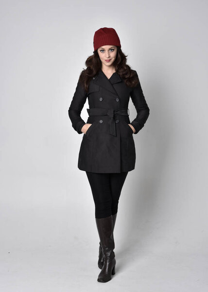 портрет красивой брюнетки в черном пальто, кожаных сапогах и красной шапочке. Стоящая поза на сером студийном фоне
.