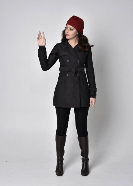 портрет красивой брюнетки в черном пальто, кожаных сапогах и красной шапочке. Стоящая поза на сером студийном фоне
.