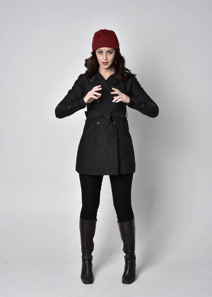портрет красивой брюнетки в черном пальто, кожаных сапогах и красной шапочке. Стоящая поза на сером студийном фоне
.