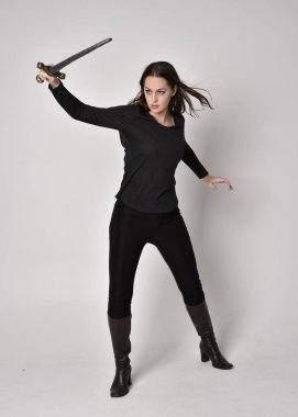 Siyah gömlekli, deri çizmeli, elinde kılıç tutan esmer bir kızın uzun bir portresi. Gri bir stüdyonun arka planında, elinde bir kılıçla dikiliyordu..
