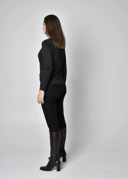 黒いシャツの革のブーツを着た美しいブルネットの女の子の完全な長さの肖像画 立ちポーズカメラから顔を背景にグレーのスタジオで — ストック写真