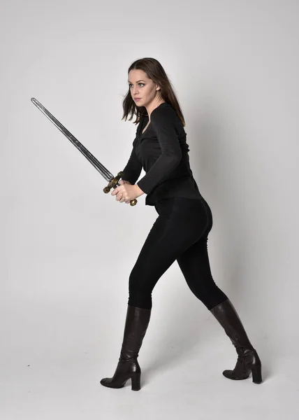 一个穿着黑色衬衫和皮靴 拿着剑的漂亮的黑发女孩的全长肖像 站在一边 举着剑 站在灰色工作室的背景下 — 图库照片