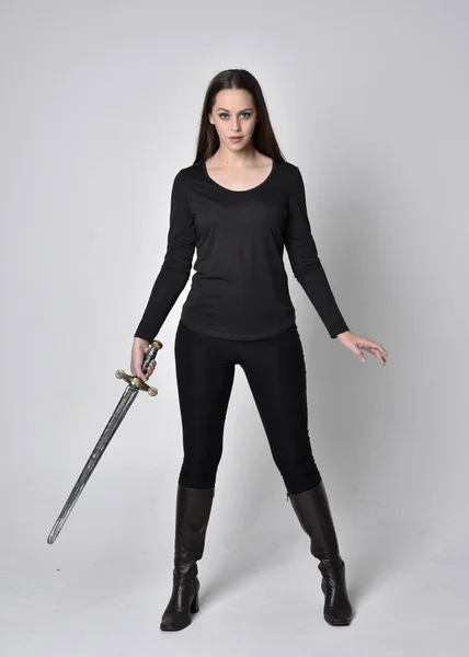 一个穿着黑色衬衫和皮靴 拿着剑的漂亮的黑发女孩的全长肖像 拿着剑 在灰色的工作室背景上 — 图库照片