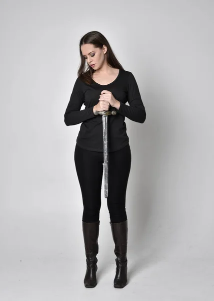 full length portrait of a pretty brunette girl wearing a black shirt and leather boots, holding a sword. Standing pose, holding a sword, on a grey studio background.