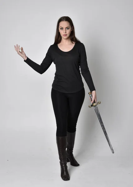 一个穿着黑色衬衫和皮靴 拿着剑的漂亮的黑发女孩的全长肖像 拿着剑 在灰色的工作室背景上 — 图库照片