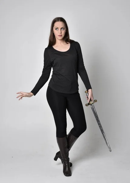 黒いシャツと革のブーツを着た美しいブルネットの女の子の完全な長さの肖像画 剣を持っています 立ちポーズ剣を持ってグレーのスタジオの背景に — ストック写真