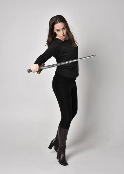 full length portrait of a pretty brunette girl wearing a black shirt and leather boots, holding a sword. Standing pose, holding a sword, on a grey studio background.