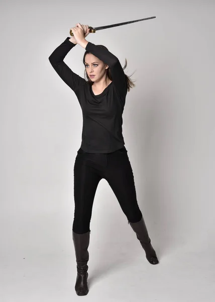 full length portrait of a pretty brunette girl wearing a black shirt and leather boots, holding a sword. Standing pose, holding a sword, on a grey studio background.