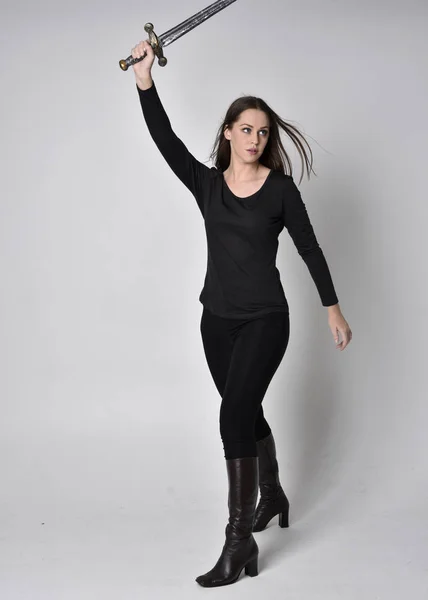 full length portrait of a pretty brunette girl wearing a black shirt and leather boots, holding a sword. Standing pose, holding a sword, on a grey studio background.
