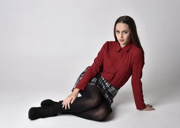 全长肖像一个漂亮的黑发女孩穿着红色衬衫和格子花裙与腿和靴子 在工作室背景上的坐姿 — 图库照片