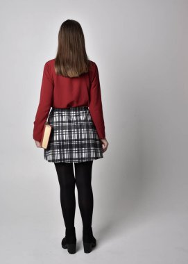 Uzun boylu, esmer, kırmızı tişörtlü, etekli, tozlu ve çizmeli güzel bir kızın portresi. Stüdyonun arka planında elinde kitap tutan poz..