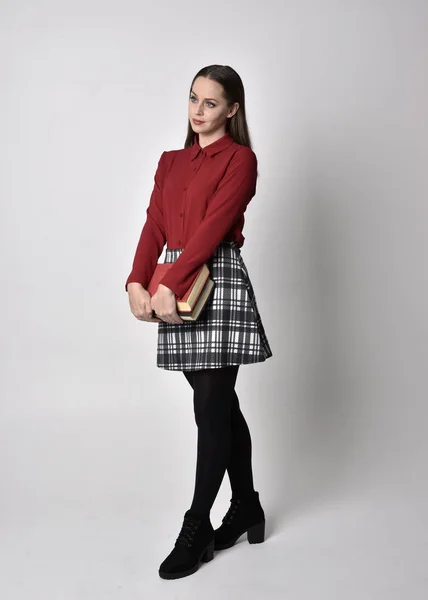 stock image full length portrait of a pretty brunette girl wearing a red shirt and plaid skirt with leggings and boots. Standing pose holding a book, on a  studio background.