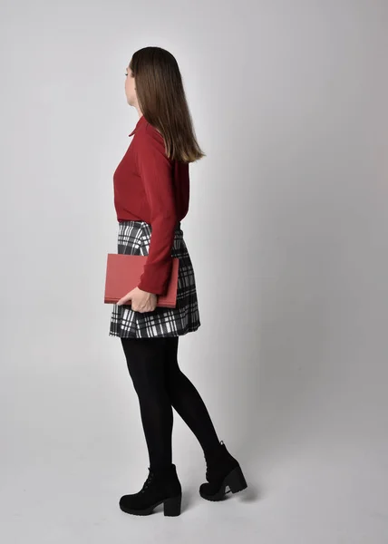 全长肖像一个漂亮的黑发女孩穿着红色衬衫和格子花裙与腿和靴子 立正举着一本书 在工作室的背景下 — 图库照片