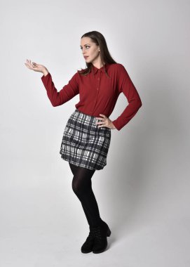 full length portrait of a pretty brunette girl wearing a red shirt and plaid skirt with leggings and boots. Standing pose with hand gesture against a  studio background. clipart