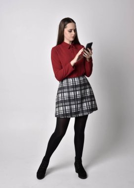 Uzun boylu, esmer, kırmızı tişörtlü, etekli, tozlu ve çizmeli güzel bir kızın portresi. Stüdyonun arka planında akıllı telefon duruşu yapıyor..