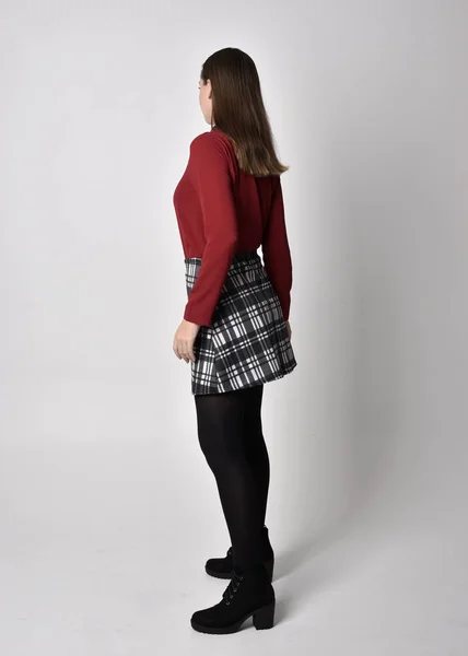 全长肖像一个漂亮的黑发女孩穿着红色衬衫和格子花裙与腿和靴子 在演播室的背景下 站在侧面的姿势 — 图库照片