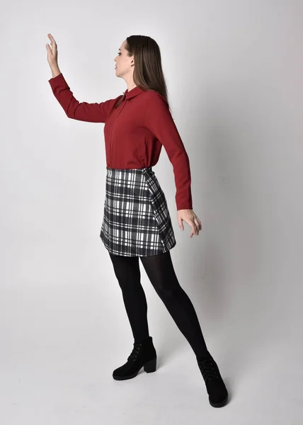 全长肖像一个漂亮的黑发女孩穿着红色衬衫和格子花裙与腿和靴子 在演播室的背景下 站在侧面的姿势 — 图库照片