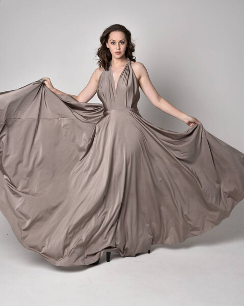 Portrait of a pretty brunette girl wearing a long silver evening gown, full length standing pose against a studio background