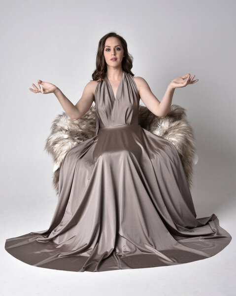 Portrait of a pretty brunette girl wearing a long silver evening gown, full length sitting pose on chair pose against a studio background.