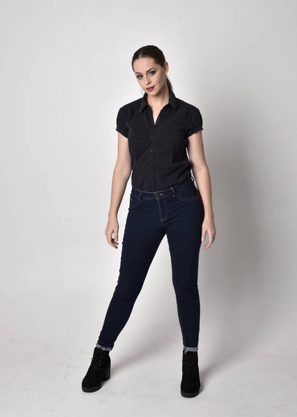 Portrait of a pretty brunette girl with ponytail, wearing jeans boots and a black blouse.  full length standing pose on a studio background.