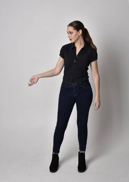 一个长着马尾辫 穿着牛仔裤靴和黑色衬衫的漂亮黑发女孩的画像 全长站立姿势在工作室的背景上 — 图库照片