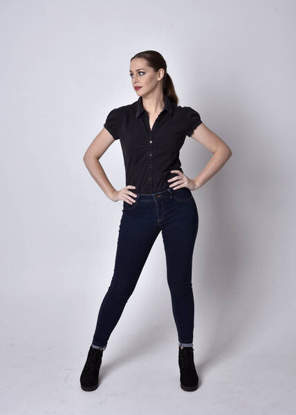 Portrait of a pretty brunette girl with ponytail, wearing jeans boots and a black blouse.  full length standing pose on a studio background.