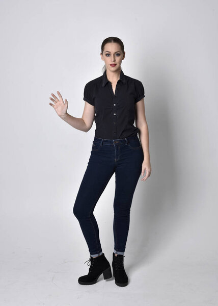 Portrait of a pretty brunette girl with ponytail, wearing jeans boots and a black blouse.  full length standing pose on a studio background.