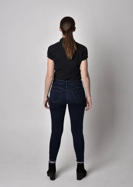 一个长着马尾辫 穿着牛仔裤靴和黑色衬衫的漂亮黑发女孩的画像 全长站立姿势 背对着摄像机在工作室的背景上 — 图库照片