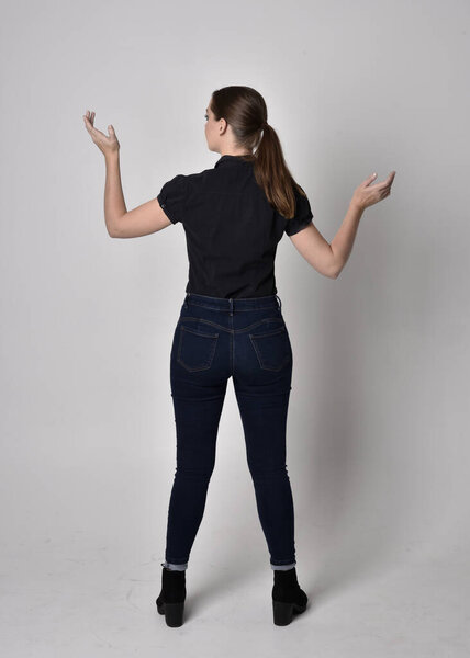 Portrait of a pretty brunette girl with ponytail, wearing jeans boots and a black blouse.  full length standing pose, with back to the camera on a studio background.