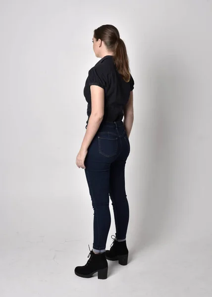 一个长着马尾辫 穿着牛仔裤靴和黑色衬衫的漂亮黑发女孩的画像 全长站立姿势 背对着摄像机在工作室的背景上 — 图库照片