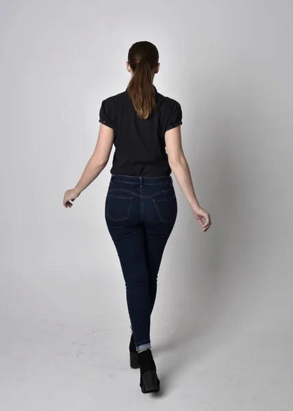 ポニーテールの可愛いブルネットの女の子の肖像 ジーンズブーツと黒いブラウスを着ています カメラに背を向けてスタジオを背に立ちポーズをとり — ストック写真
