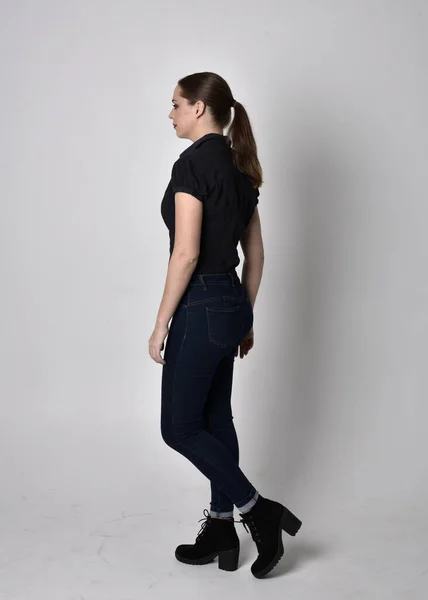 一个长着马尾辫 穿着牛仔裤靴和黑色衬衫的漂亮黑发女孩的画像 全长站立姿势 在工作室背景上 — 图库照片