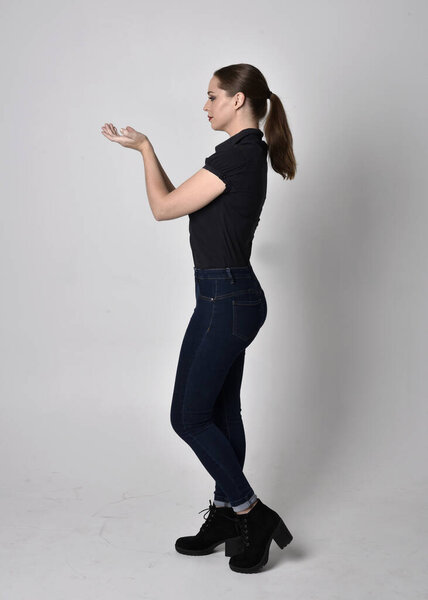 Portrait of a pretty brunette girl with ponytail, wearing jeans boots and a black blouse.  full length standing pose in side profile,  on a studio background.