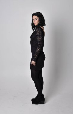 Siyah dantelli elbise ve çizme giyen koyu saçlı gotik bir kızın portresi. Stüdyo arka planında tam boy ayakta poz.