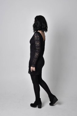 Siyah dantelli elbise ve çizme giyen koyu saçlı gotik bir kızın portresi. Stüdyo arka planında tam boy ayakta poz.