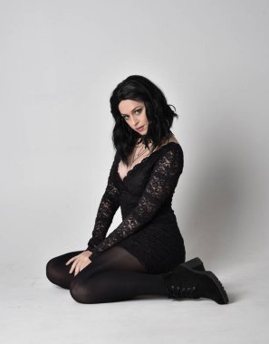 Siyah dantelli elbise ve çizme giyen koyu saçlı gotik bir kızın portresi. Tam boy oturma pozisyonu, stüdyo arka planında..