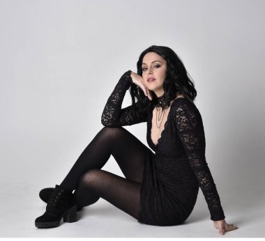 Siyah dantelli elbise ve çizme giyen koyu saçlı gotik bir kızın portresi. Tam boy oturma pozisyonu, stüdyo arka planında..