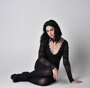Siyah dantelli elbise ve çizme giyen koyu saçlı gotik bir kızın portresi. Tam boy oturma pozisyonu, stüdyo arka planında..