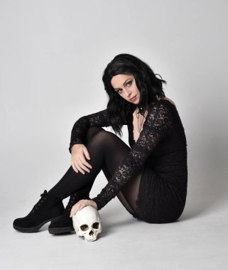 Siyah dantelli elbise ve çizme giyen koyu saçlı gotik bir kızın portresi. Tam uzunlukta oturma pozisyonu ve bir insan kafatasını stüdyo arka planında tutma.