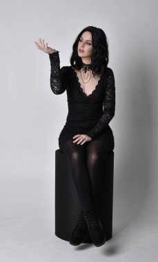 Siyah dantelli elbise ve çizme giyen koyu saçlı gotik bir kızın portresi. Stüdyo arka planında tam boy oturma pozu.