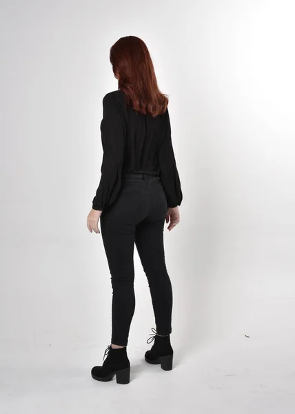 黒いジーンズ ブーツ ブラウスを着た赤い髪のきれいな女の子の肖像画 カメラに背を向けてスタジオの背景に立ち — ストック写真