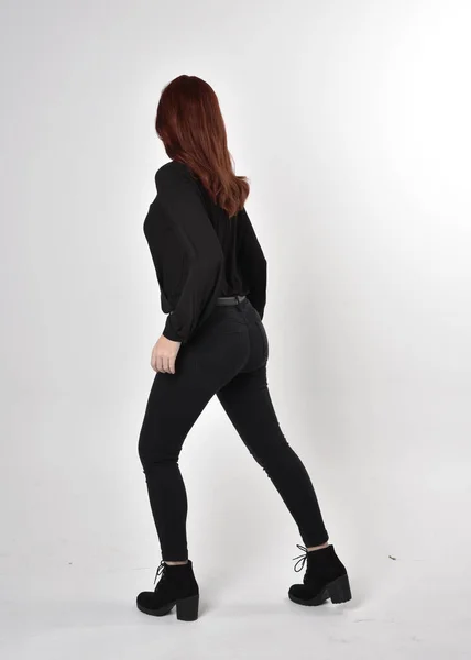 一个红头发 穿着黑色牛仔裤 靴子和衬衫的漂亮女孩的画像 全长站立姿势在工作室的背景上 背对着相机 — 图库照片