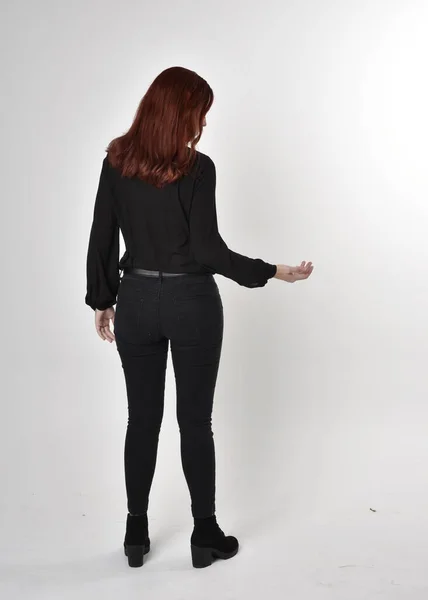 一个红头发 穿着黑色牛仔裤 靴子和衬衫的漂亮女孩的画像 全长站立姿势在工作室的背景上 背对着相机 — 图库照片