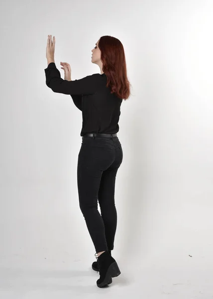 黒いジーンズ ブーツ ブラウスを着た赤い髪のきれいな女の子の肖像画 カメラに背を向けてスタジオの背景に立ち — ストック写真