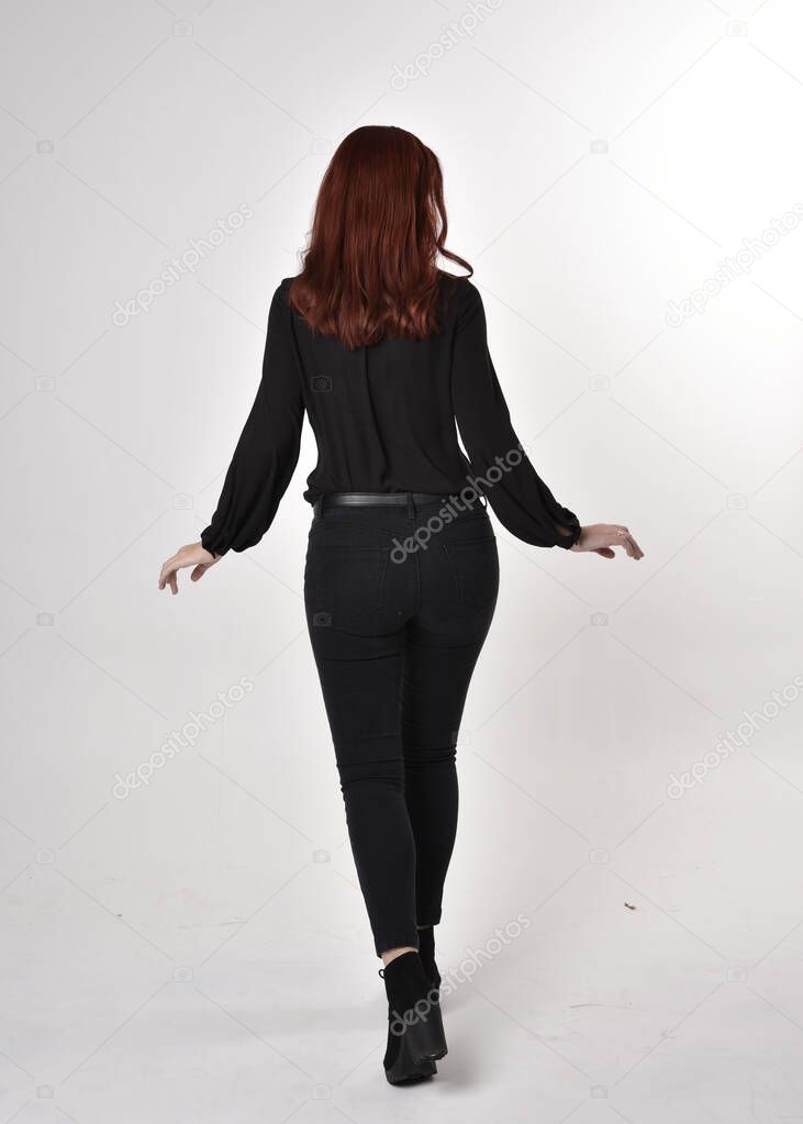 Portrait of a pretty girl with red hair wearing black jeans, boots and a blouse.  full length standing pose on a studio background, with back to the camera.
