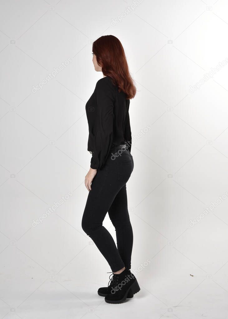 Portrait of a pretty girl with red hair wearing black jeans, boots and a blouse.  full length standing pose on a studio background, with back to the camera.