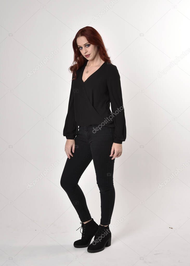 Portrait of a pretty girl with red hair wearing black jeans, boots and a blouse.  full length standing pose, facing the camera, on a studio background.